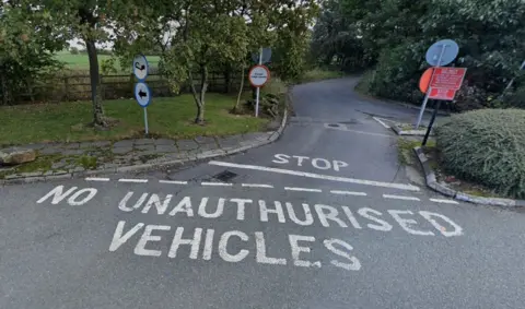 Google A tree-lined country lane winds away around a bend with a junction in the foreground that has "no unauthorised vehicles" written on the tarmac.