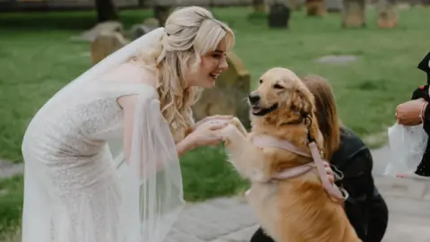 Harper Scott Jenn Nimmo-Smith - a woman with long blonde hair and wearing a white wedding dress - holds the paws of her golden retriever dog Scout in her hands. Scout is standing up on her back paws.