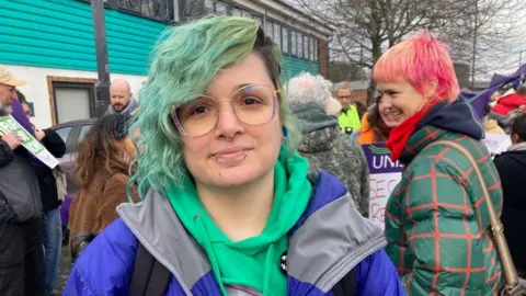 A young woman with green hair, glasses and piercings, wearing a blue jacket, stands on the picket line outside the Second Step HQ in a crowd of striking workers.