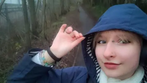 A selfie image of Amelka from the shoulders up, she is wearing a navy rain coat with the hood up and a white scarf. She is on a gloomy walking path, it seems to have been raining and it's cold.