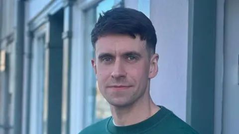 Eóin McAlonan, a man with short, dark hair, looking to the camera on a street in Ballycastle. He is wearing a green crew-neck sweatshirt. There are steel barriers behind him and a church with a clock tower in the background.