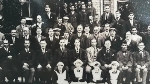 Kitchener's Guest Home A black and white photo of the mostly male staff at Lord Kitchener's Memorial Home in August 1924. The staff are sitting in three rows, with four women, in servants' uniform of white cap and white apron, sitting on the ground in front of the men. The men are in suits, some wearing flat caps. Behind them staff are more men, sitting and standing, with a group clustered in the doorway of the building they are in front of. 