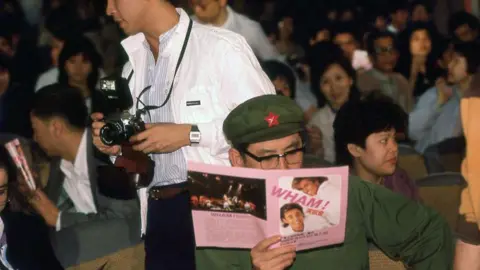 Andrew Ridgeley, Big Geoff Overseas Limited/Robobuild Limited/George Michael Entertainment The audience of a Wham! performance in China. One man, dressed in a green uniform, is reading a pink tour programme.