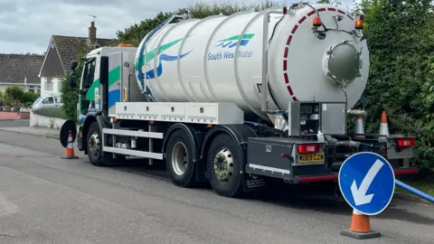 A South West Water lorry is parked in a residential area. There us a cone with an arrow sign in front and behind it and a wide hose pipe leading from the lorry out of shot.