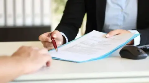 A hand is holding a piece of paper and a red pen on a desk. 