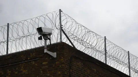 A CCTV camera sits at the top of one of the outside walls of HMP Pentonville in London. Above it, the brick wall is lined with barbed wire.