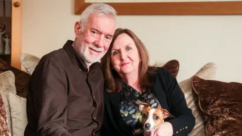 A man, a woman and a dog sit together on a sofa to pose for a photo. The man is wearing a brown top and green trousers. The woman is wearing a black cardigan and atop decorated with petals and jeans. The small brown and white dog, who is a jack russell, is sitting on her lap. The man has his arm around the shoulders of the woman and the woman's hands are on the dog.