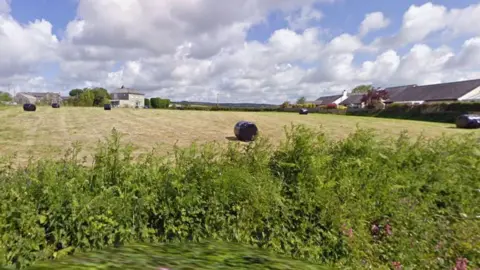 A field in St Dominick. In the foreground is a lush green hedge. Beyond the hedge is a large field which contains six wrapped bales of hay. The field is surrounded by more hedges around the perimeter and a couple of houses at the back and right hand side. It's a cloudy day.