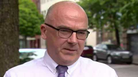 Patrick Mulholland is standing near a street with a number of parked cars and trees. He is wearing black glasses with a white shirt and purple tie.