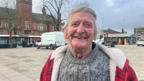 Alan Braddock with white hair wearing a grey woolly jumper and red and black fleece smiling in Hyde town centre