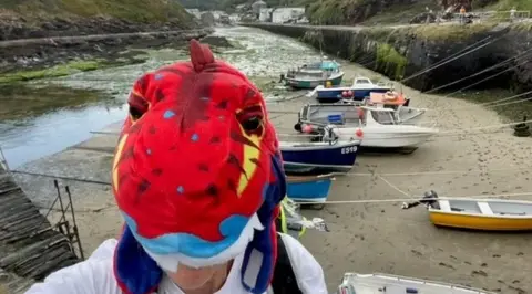 Woman wearing a colourful hat which looks like a dinosaur with boats and sand in the background.