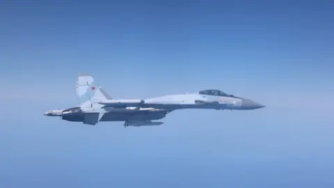 A grey Russian fighter jet is flying against a clear blue sky.