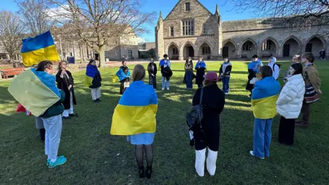 A number of people, many draped in blue and yellow Ukrainian flags stand in a circle on grass in front of university buildings 