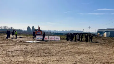 Luke Deal/BBC A large group of people stand to the right of the image. A digger rests in the middle behind two white signs. A camera crew interview some people to the left of the image. 