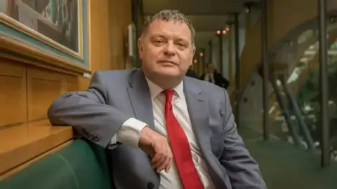 Mike Amesbury sat wearing a suit with a red tie leaning on a green leather seat