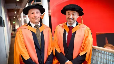 Redpix Nikolai Foster pictured on the left and Chris Stafford right. Bot men are wearing orange gowns and black caps. They are also both wearing suits and ties. 