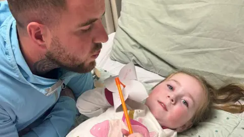 Sian Kennedy Sienna lying in a hospital bed, looking at the camera and holding a paper windmill toy, while her father Jack Gibson, wearing a blue hooded top, blows at the windmill.