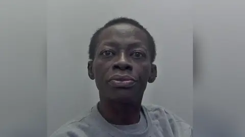 A police mugshot of a woman in a grey jumper with shaved hair. 