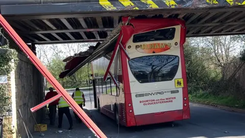 A Pulhams double decker bus is lodged beneath a railway bridge on a country road, with its roof squashed and partly ripped off. There are men in hi-vis tabards stood around it and clear up items, such as brooms, have been propped up against the bridge's pillar.