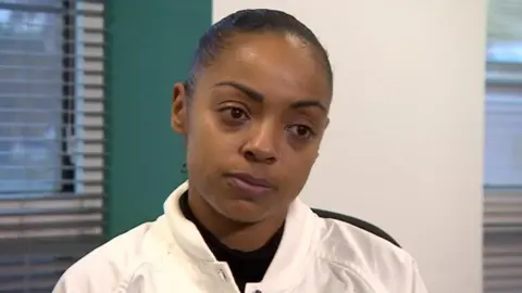 Tiffany Williams wearing a white windbreaker jacket and sitting in a room looking just off to the right of the camera. She has dark hair which is pulled back into a ponytail and a silver nose ring. She has a solemn expression.