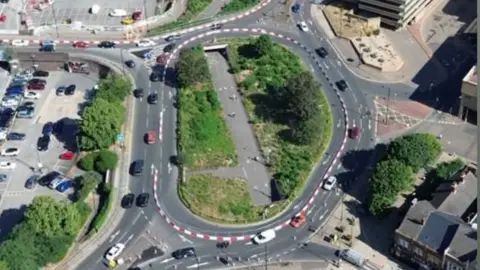 An aerial view of a roundabout with cars driving round it. The roofs of buildings and a car park dot the landscape along with a few with areas of trees and grass. 