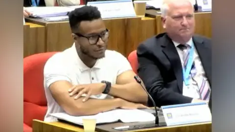 Lambeth Council A screen shot of a council meeting - a councillor sits at a desk and is wearing a white T-shirt and glasses sits with his arms folded over some paperwork. There is a sign in front of him that reads Cllr Andre Bourne. Beside sits another man in a dark suit, white shirts and striped tie. There are other people sat at similar desks in the background. 