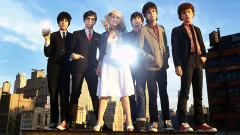 Members of Blondie stand together on a rooftop in New York City in 1978, with Debbie Harry in the centre and a camera flash reflecting off her white dress.
