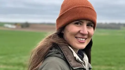 A woman in a beanie hat and a green coat stands in a field. She is smiling at the camera.
