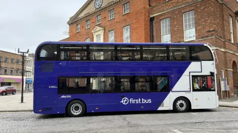 BBC A first bus with blue and white branding. It is in the centre of Taunton outside an old victorian bricked building. 