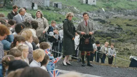 Getty Images Diana is wearing a long, green raincoat. She is standing next to Charles who is wearing a kilt and a tweed jacket and waist coat. There islanders gathered around them.