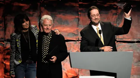 Getty Images Chrissie Hynde with her arm around Billy Steinberg, watching Kelly standing at a podium and holding an award aloft