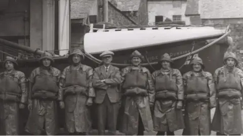 National Historic Ships Registry A black and white image of men standing in front of the vessel. The men are lined up in a row and are covering the vessel's name. They are wearing hats, life jackets and long coats.