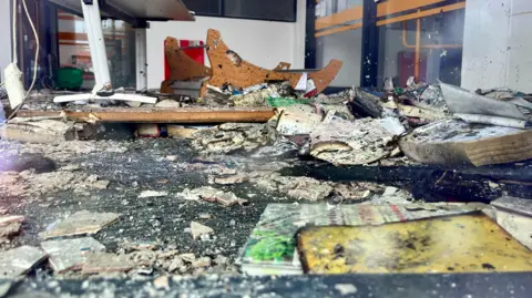 Rubble is smashed on the floor, with damaged books, hanging wires, a wooden bookshelf on its side and a plank of wood with nails sticking out.  