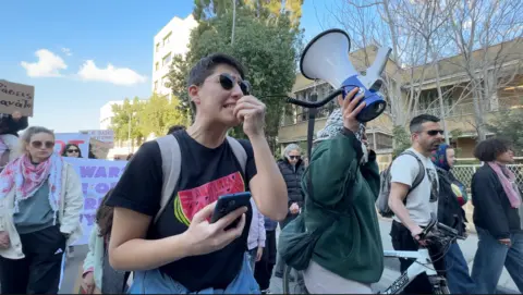 Jessica Parker/BBC A person in a black T-shirt with a watermelon on the front speaking expressively into a megaphone while another person holds the receiver, amid a crowd of protesters walking together through the street with some holding up posters. 