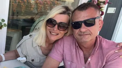 Julia Buzar A man and woman wearing dark glasses sit at a cafe table. The blonde woman has her arm around the man who is wearing a pink short-sleeved shirt. 