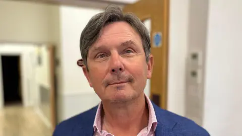BBC Deputy Andy Sloan. He has short dark hair and is wearing a pink shirt and a blue suit jacket