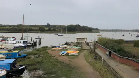 Vikki Irwin/BBC A selection of boats moor on the side of a river. In the distance boats are also moored on the river. On the right a man and woman are walking on a path.