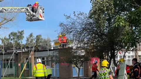 Cambridgeshire Fire and Rescue A firefighter in a crane lift above a low-lying school building with two firefighters on the roof and six others milling around below after a fire