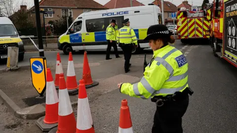 A female police officer stands on a road beside traffic cones with two police vans, two fire engines and two other male police officers behind her