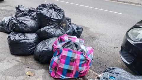Basmin Khan Roughly ten black bin bags piled up on a street, some on the pavement and some spilling onto the road.