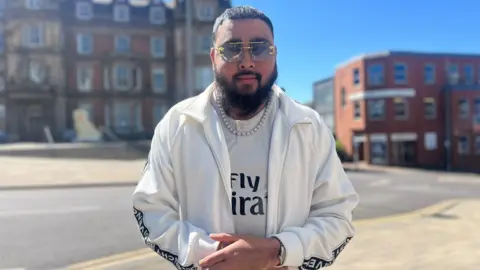A man stands on a street on a sunny day. He has close-cut black hair, a black beard, and sunglasses. He is wearing a white tracksuit top, a white football shirt, and a silver necklace. Red brick buildings are behind him.