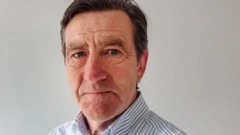 A closeup of Derek Flint, a middle-aged man with dark hair who is wearing a striped shirt and looking sternly at the camera.