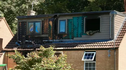 Martin Giles/BBC Three sets of fire-damaged windows can be seen as part of a loft extension on the roof of a sand coloured brick house, with trees seen in the background.
