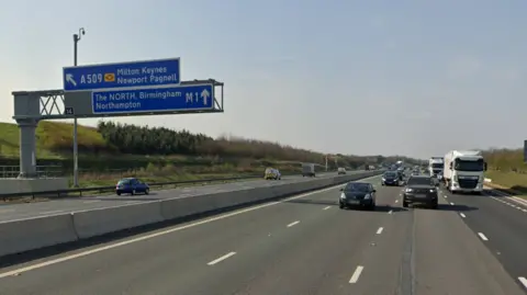 A Google Maps street view image of the M1 between junctions 14 and 15. Vehicles are travelling on the road in both directions and a blue sign directing drivers towards Milton Keynes and the North can be seen on the left. 