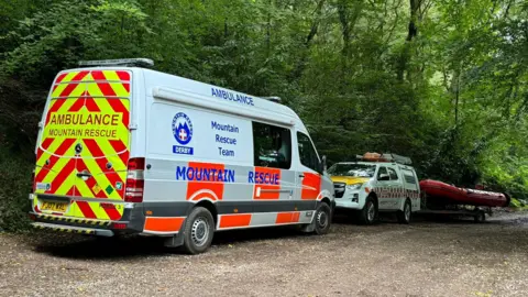 A white mountain rescue van from Derby in front of a smaller van with a search boat behind it, parked on a woodland path with trees in the background.