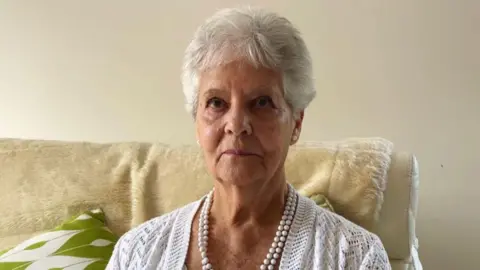 A recent picture of Ann, she has short grey hear and is wearing a white cardigan and a pearl necklace. 