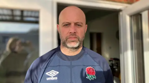 James Richardson stands outside his house, looking at the camera. He is wearing a navy blue t-shirt. He is balding and has a grayish-brown beard and blue eyes.