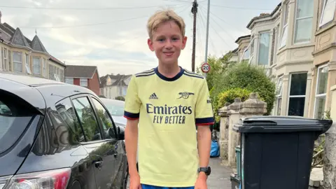 George stands in the street and smiles at the camera. He is wearing a light yellow Arsenal shirt. 