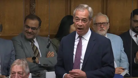 Nigel Farage stands speaking in the House of Commons, wearing a dark suit, white shirt, and patterned tie. He is surrounded by other members of Parliament in a packed Chamber