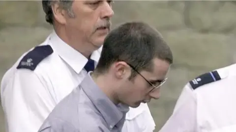 PA McKilligan being escorted by two police officers in white shirts. McKilligan has short brown hair, is wearing glasses and a blue shirt.